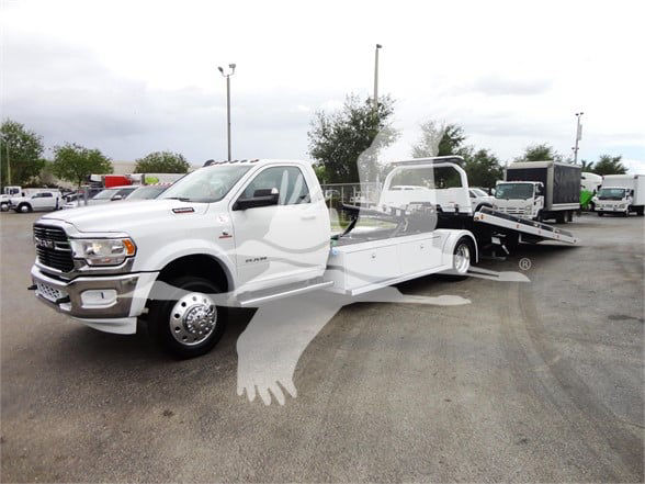 Truck in a dealership lot: white 2023 Ram 5500 HD SLT tow truck with flatbed, front chrome bumper, dual rear wheels visible, dealership background.