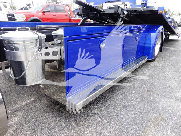 Blue Peterbilt 337 tow truck deck side with chrome fuel tank and wheel well; reflective surface; other vehicles in background.