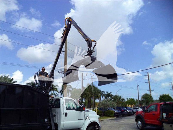 Front-left view of a 2023 Ford F650 SD grapple truck with a raised hydraulic arm, workers in a bucket, and a white cab in a dealership lot.