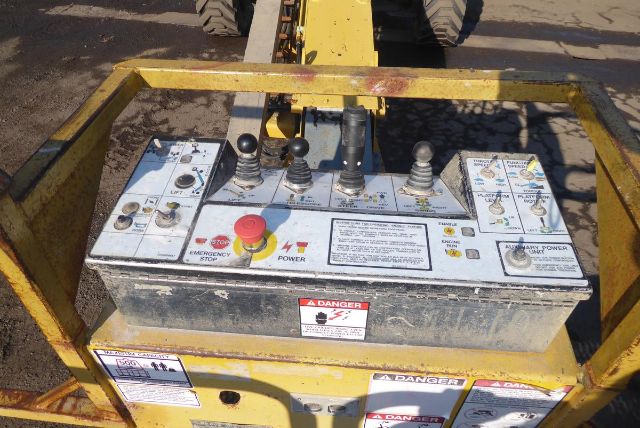 Control panel of a Grove T60 crane shows multiple levers, buttons, and labels. Red emergency stop button, power switch, various warning stickers, and instruction panels on a metal console.