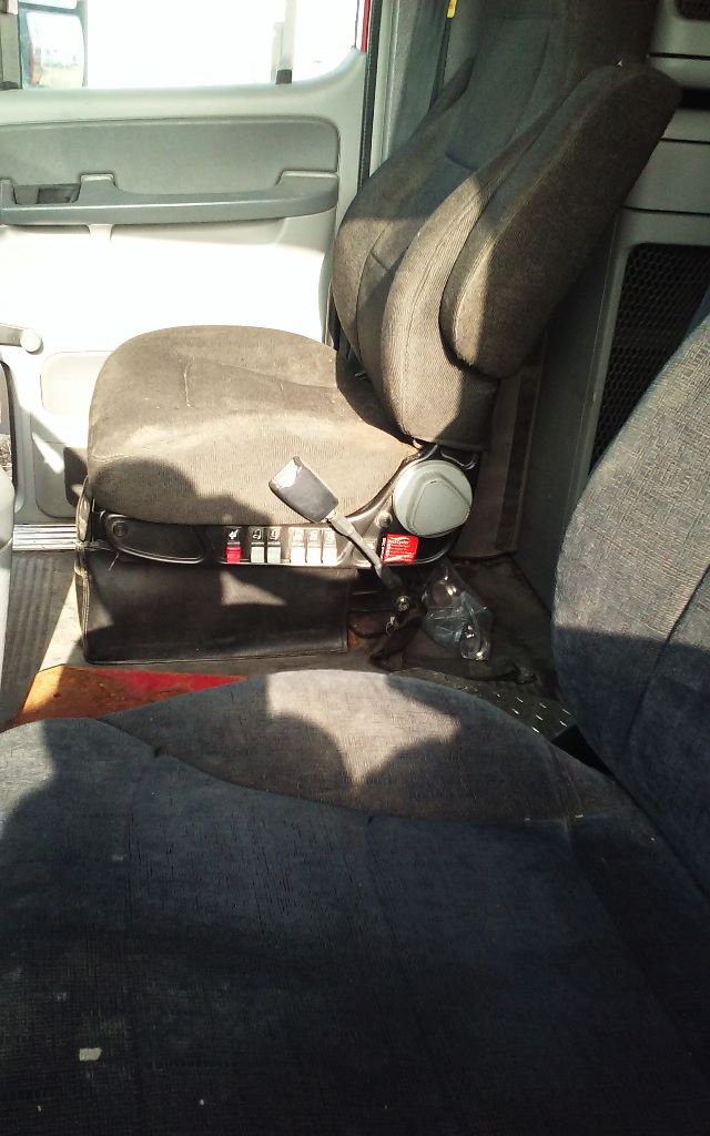Cab interior of a Freightliner truck showing driver seat with controls and armrest, gray door panel, fabric seats, and dashboard edge.