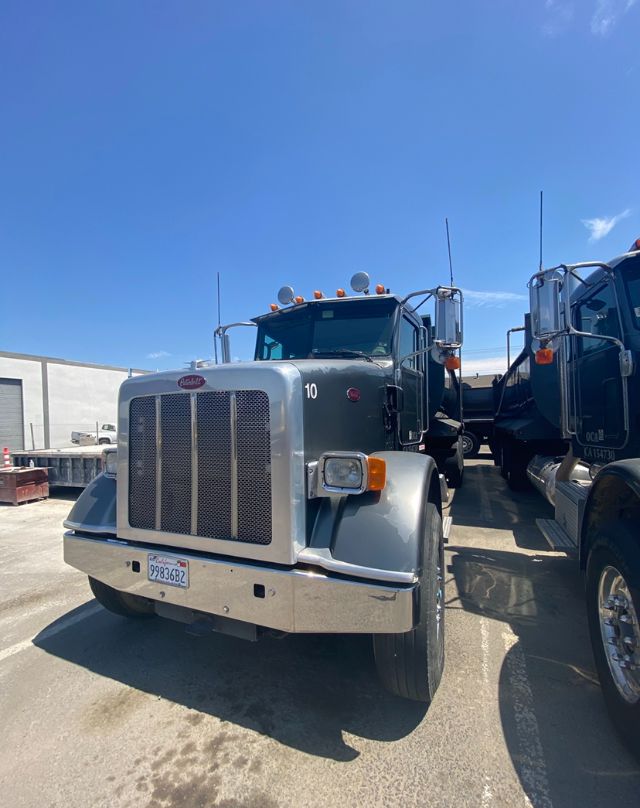 Peterbilt dump truck front view showing large grille, chrome bumper, rectangular headlights, and side mirrors with stacked vehicles in background.