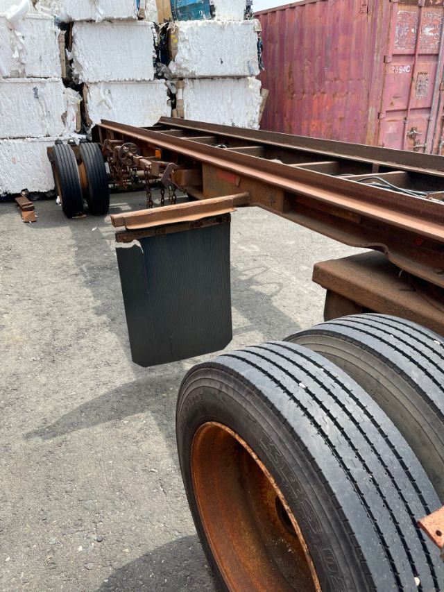 Trailer frame with rusted rails and dual rear axles; black heavy fenders and tires in foreground; stacked white blocks and red container in background.