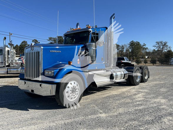 Kenworth W900L conventional daycab truck in blue, front grille with Kenworth badge, dual exhaust stacks, large chrome bumper, white wheels, gravel lot.