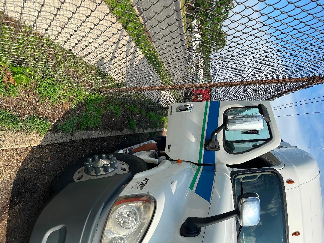 Truck cab of a 2011 Freightliner Cascadia with blue and green striping, front wheel and mirror visible, chain-link fence in background.