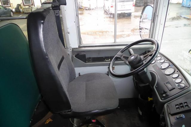 Bus cockpit with steering wheel and gray driver seat, dashboard gauges, and windows showing outside yard.
