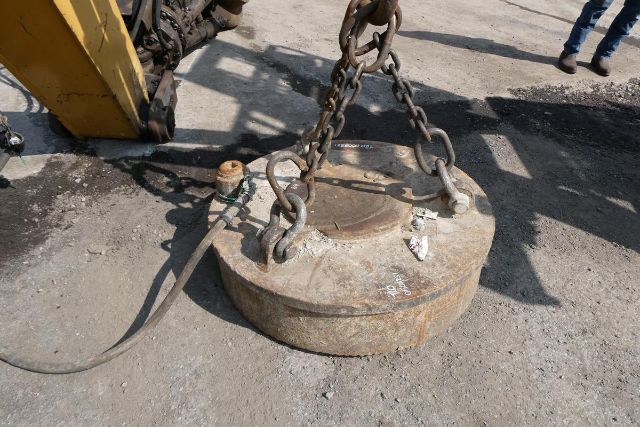 Circular metal weight/detector with chains and hooks attached to a forklift or crane, lifting eye bolts and a rope; rusty metal surface and concrete ground.