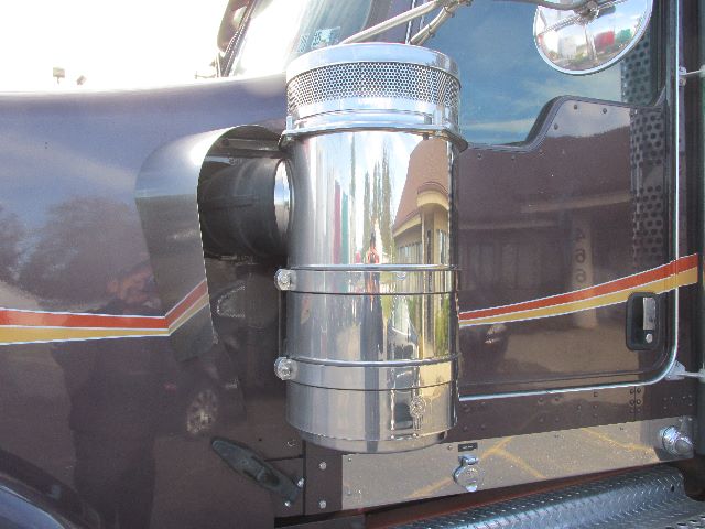 Close-up of a Kenworth W900 truck cab side showing a polished cylindrical air filter housing with chrome bands and mounting clamps, part of the engine bay area.