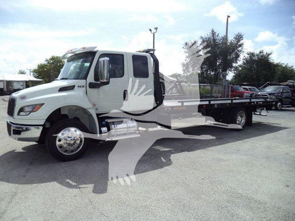 International MV tow truck with flatbed, cab door open, showing side profile and wheel, dealership lot in the background.