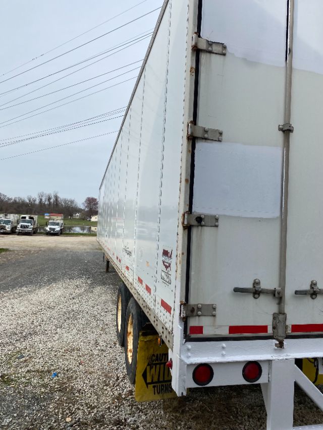Trailer side view showing white Wabash Duraplate trailer with rear doors, double axles, yellow wheel chocks or mud flaps, and red reflective strips.