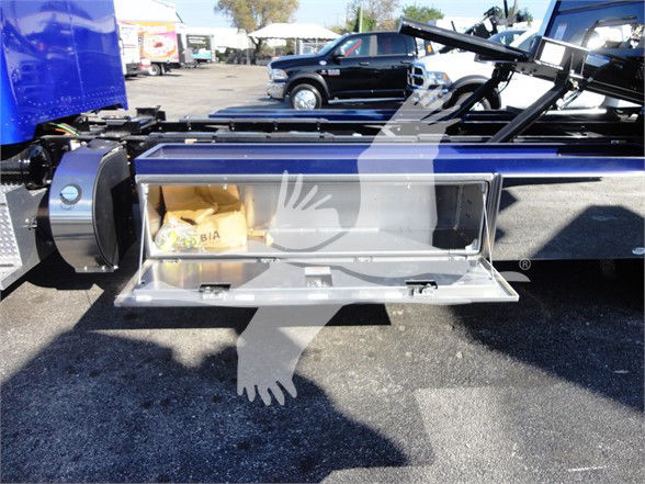 Tow truck chassis with a hinged metal storage compartment open, showing a tan paper bag-like item inside. Background shows other trucks and a parking lot.