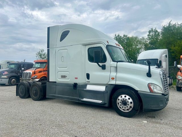 White Freightliner Cascadia semi-truck sleeper cab side view showing front wheel, steps, and tall aerodynamic roof in a lot with other trucks.