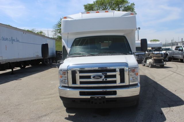 Front view of a 2025 Ford E450 Shuttle Bus with large chrome grille, side mirrors, and elevated roofbox in a dealership lot.
