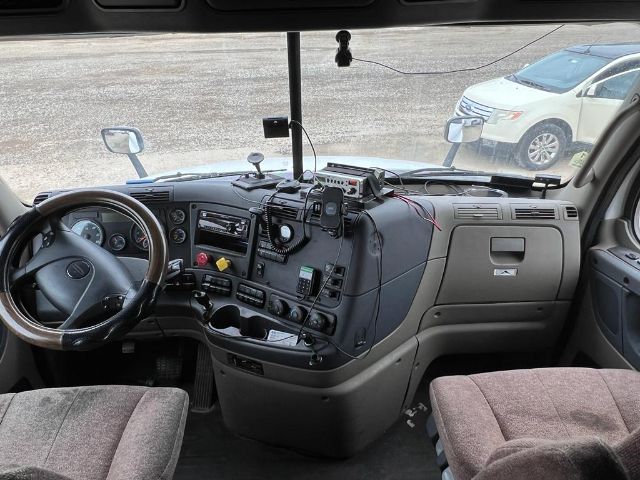 Cab interior of a Freightliner Cascadia truck showing steering wheel, dashboard with gauges, radio, and various mounted devices.