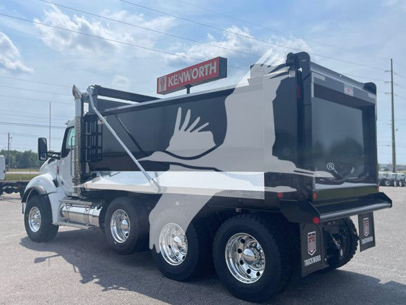 Kenworth T880 dump truck in a dealership lot with large Kenworth logo on a sign; side view shows three axles, dump bed, chrome wheels, and branding on the rear mud flaps.