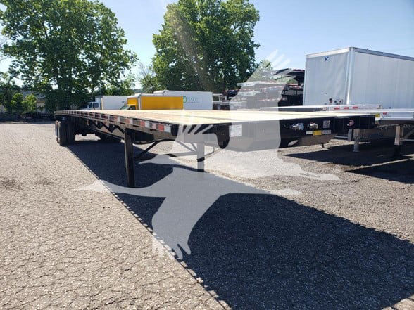 Flatbed trailer showing a long steel deck with support legs, side rails, and some equipment on the front; trees and vehicles in background.