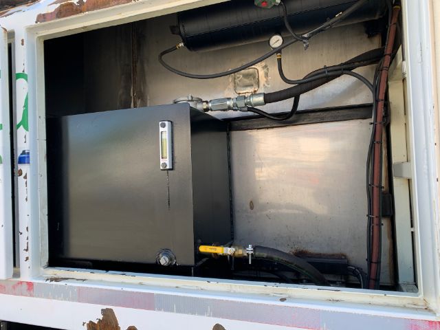 Open rear compartment of a garbage truck showing a large dark metal cabinet with a small rectangular window, hoses, and a yellow-handled valve; surrounding metal panels and wiring.
