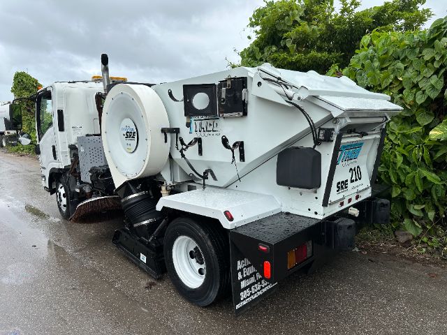 Isuzu NPR-HD sweeper truck with white body and rear vacuum/sweeper equipment; spare tire mounted on side; various hoses and panels; the back shows SRE and 210 markings.