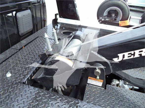 Tow truck bed with diamond-plate surface, mounting hardware, and a spare wheel. Visible text includes 'JER' on the side of the truck bed and a small orange warning label near a hitch area.