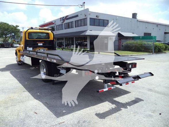Flatbed tow truck with a yellow cab and long metal flatbed in a dealership lot.