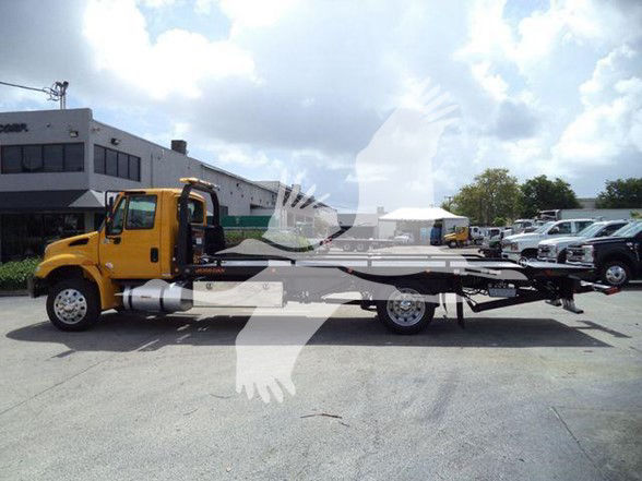 Image of a yellow tow truck chassis with a long flatbed, parked in a lot with other vehicles in the background.