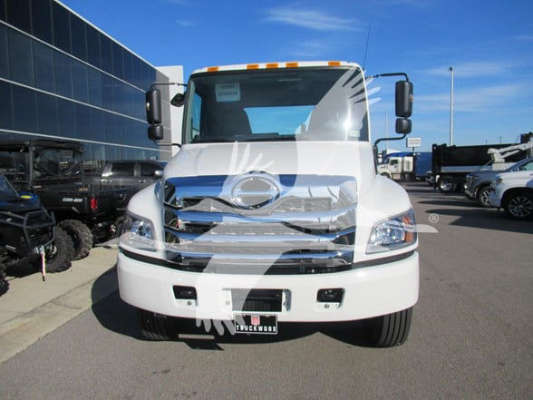 Front view of a white 2026 Hino L6 cab and chassis truck with damaged front bumper and chrome grille. Side mirrors visible, parking lot background.