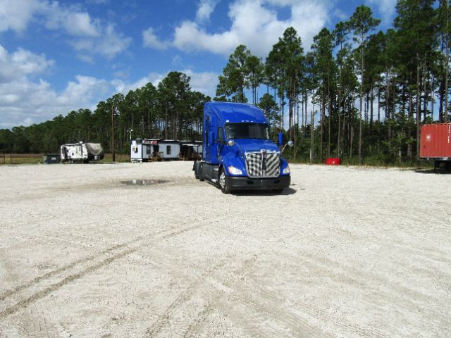 Kenworth T680 blue truck in a gravel lot with trees in background.