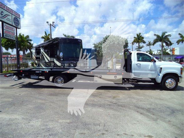 2023 Chevrolet Silverado 6500HD tow truck with flatbed, white cab, and black flatbed in a lot; palm trees and dealership signs in background.