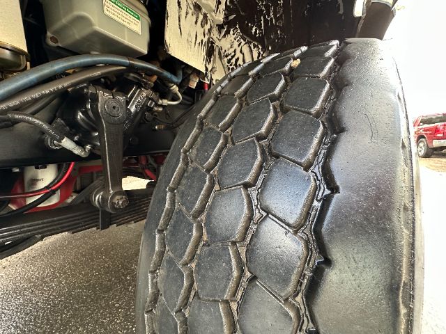 Front left tire of a 2011 Freightliner M2 truck showing dual front axle components and part of engine area.