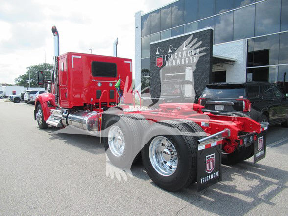 Kenworth W990 conventional sleeper truck on lot, red tractor with chrome fuel tanks and dual rear axles, showing rear view with factory decals.