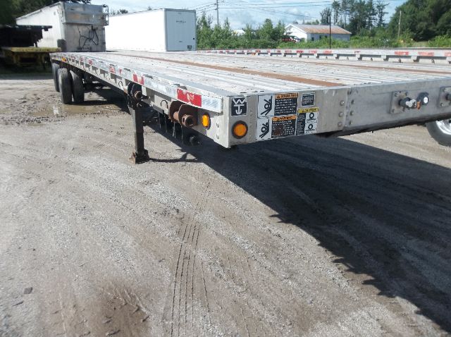 Flatbed trailer showing side rails, jack stand, and warning labels near the front. Various stickers with caution icons and reflective tape along the edge.
