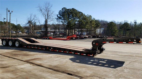 Fontaine lowboy trailer in railway yard showing long flatbed with roller paver, black gooseneck hitch, multiple axles, and safety reflectors along the sides.