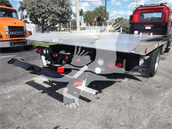 Flatbed tow truck bed with rear ramps and hitch assembly. Visible text: none clearly legible on the bed; background shows other trucks.