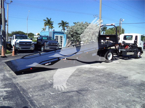 Flatbed tow truck with a long black lowered ramp on a tow truck trailer, parked lot with palm trees and other trucks in background.
