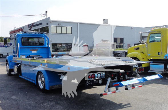 Blue Peterbilt 337 tow truck with a flatbed, seen from rear-left side in a dealership lot.