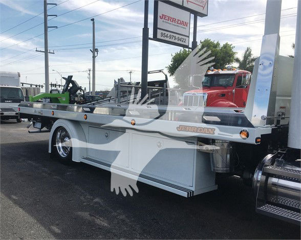 Peterbilt 337 tow truck flatbed side view showing storage compartments and lettering 'JERR-DAN' on deck, dealership lot in background.