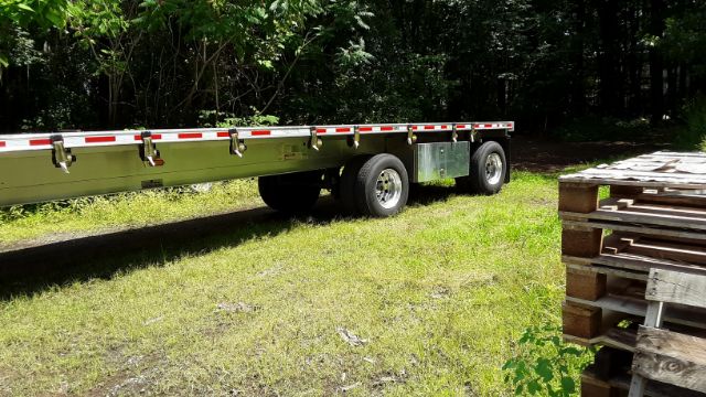 Flatbed trailer in a grassy area with stacked wooden pallets on the side, two rear axles visible and reflective safety tape along the edge.