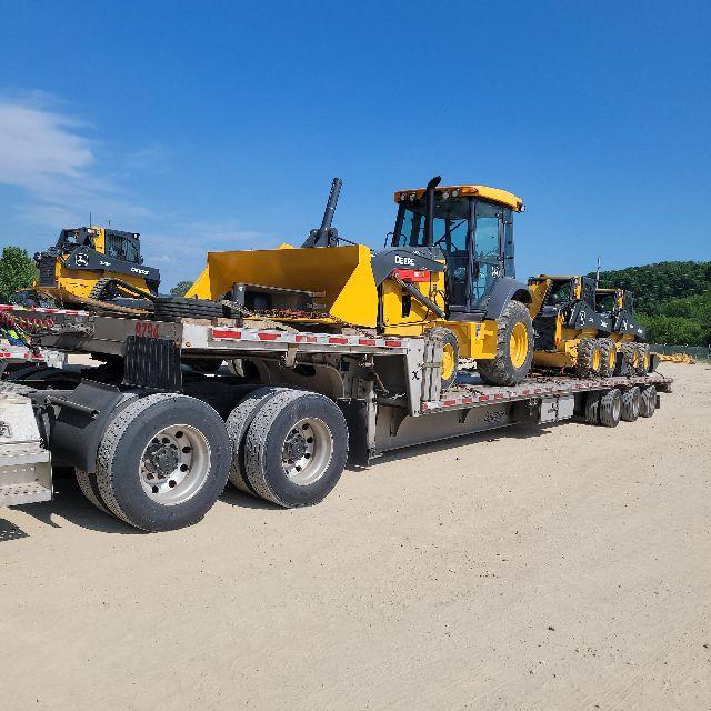 Truck trailer carrying multiple yellow John Deere machines with a drop deck trailer on a sandy lot.