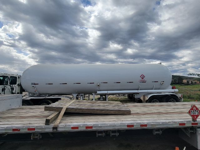 A large white cylindrical propane/chemical tanker trailer on a flatbed truck. Side markings include hazard placards and small text, with a cloudy sky above. Wooden planks on the trailer bed.