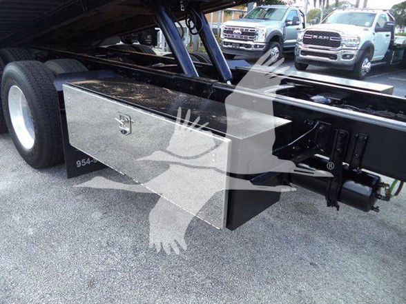 Truck chassis with metal toolbox mounted beneath the cab, showing a rectangular steel storage box and hydraulic lines near the frame of a Freightliner Business Class M2 106.