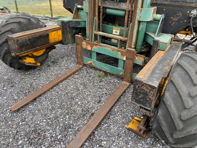 Kesmac F5000 forklift forks and front carriage showing steel forks attached to green lift mechanism and large rear tire in gravel surface.