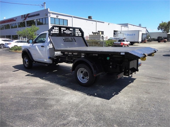 Truck bed with a flatbed/dump style, Ram 5500 HD flatbed with a metal grated cab back and a heavy-duty tow hooklift, no visible logos on side.