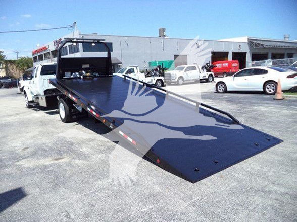 Flatbed tow truck with a long black steel bed, front cab visible in background, a white pickup in the distance, and a clear blue sky.