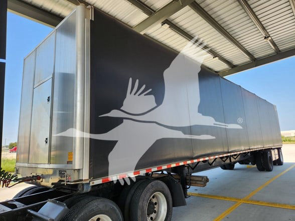 Curtainside trailer with large white seagull logo on dark side panel. 2022 Transcraft trailer under a metal roof, rear doors visible, undercarriage with wheels and axles.