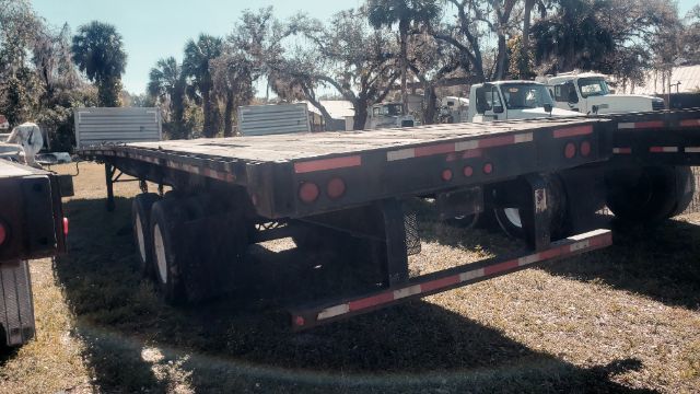 Flatbed trailer with dual rear axles and long deck, visible taillights and rear bumper; no legible branding or text on the trailer.