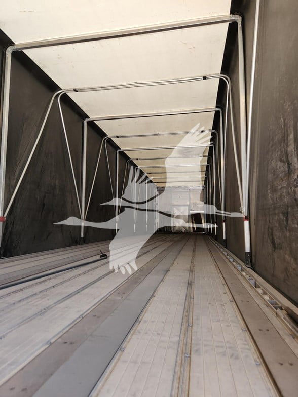 Curtainside trailer interior with side rails and overhead bows, empty long cargo deck extending to back, metal floor with track grooves.