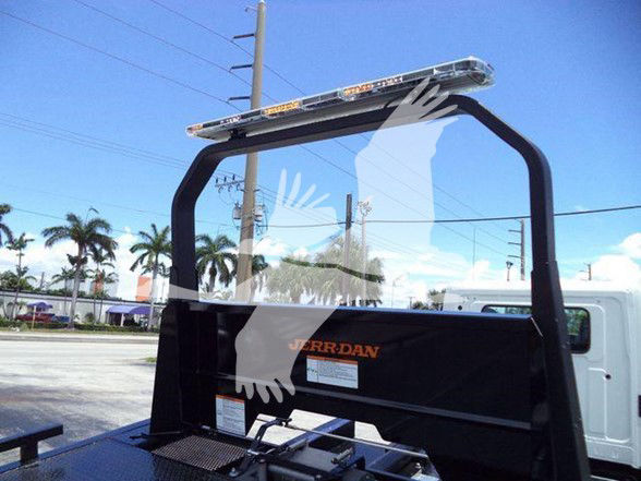 Tow truck equipment with a black metal rack and ladder-like frame; a clear glass or plastic screen in front; an orange label and white stickers on the panel; palm trees and power lines in the background.