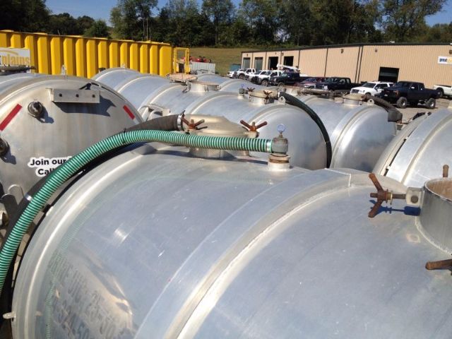 Storage tanks on a lot with a green flexible hose connected to a fitting on a metal tank; other similar tanks visible in the background.