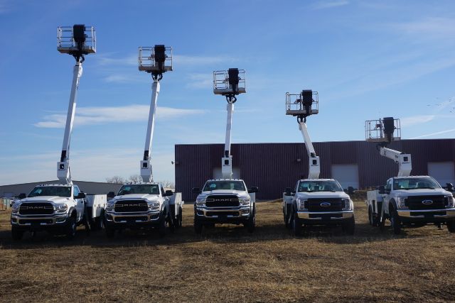 Five white Ford F-550 trucks with Manitex A62 aerial lifts raised in a row, each on a hillside with a dark barn-like building in the background.