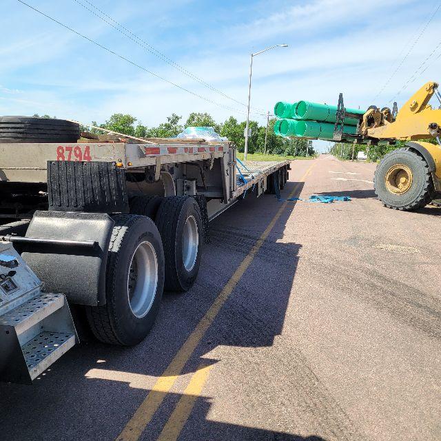 Semi-trailer with 53’ flatbed showing wheel area, number 8794 on the side, and a loader lifting green pipes in the background.
